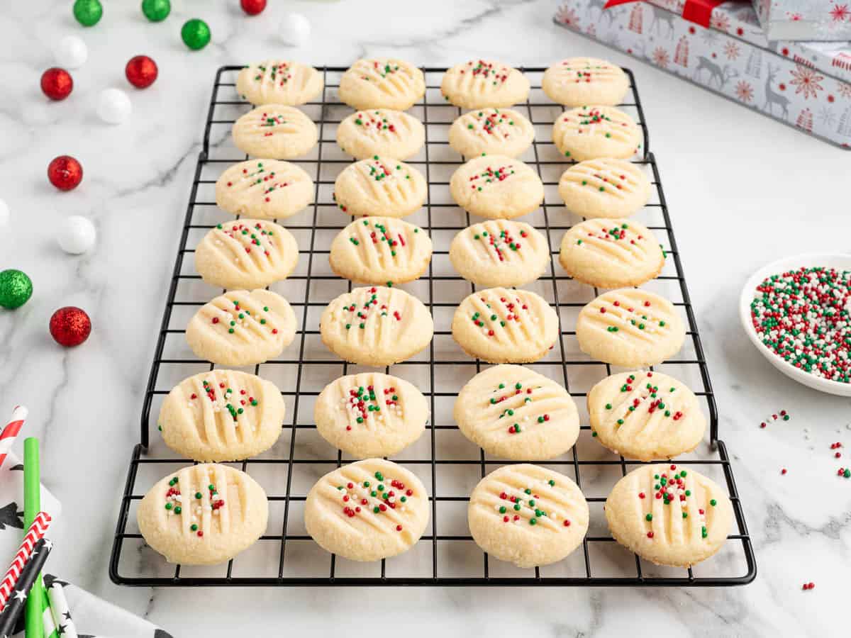 A cooling rack holds rows of round, festive Christmas Shortbread Cookies topped with red, green, and white sprinkles. Holiday decorations and a gift box surround the cookies on a white marble surface.