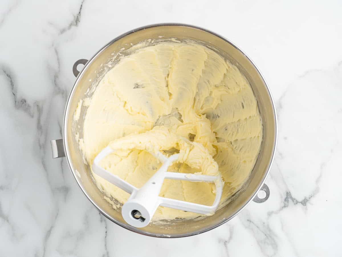 Pale whipped butter for Christmas shortbread cookies in a metal mixing bowl, with the paddle attachment resting in it on a white marble surface.