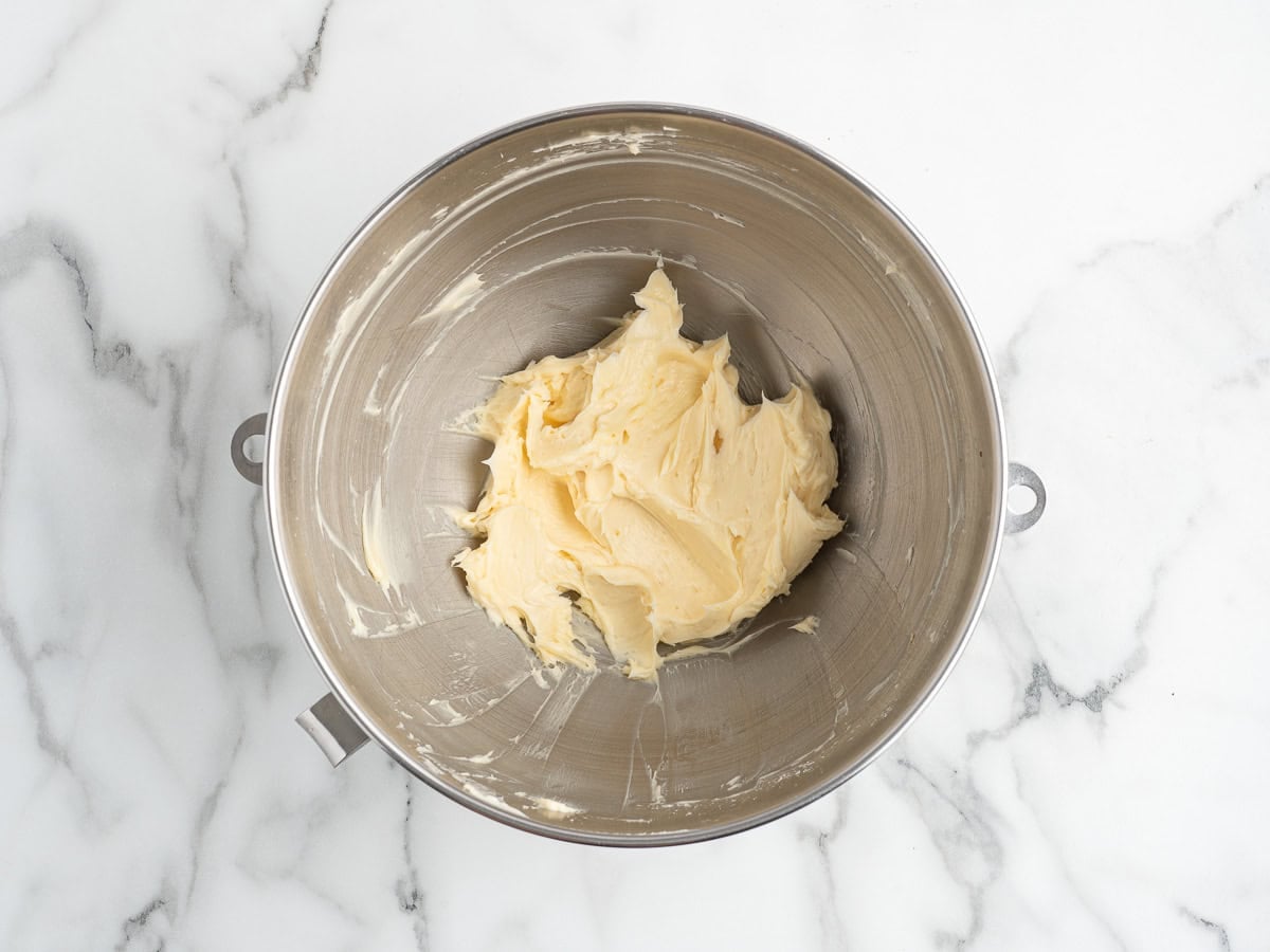 The whipped butter and powdered sugar for Christmas shortbread cookies, pale and fluffy in a metal mixing bowl on a white marble surface.