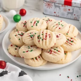 A plate of round Christmas Shortbread Cookies topped with red and green sprinkles, surrounded by holiday decorations, a bowl of sprinkles, wrapped gifts, and a glass of milk.