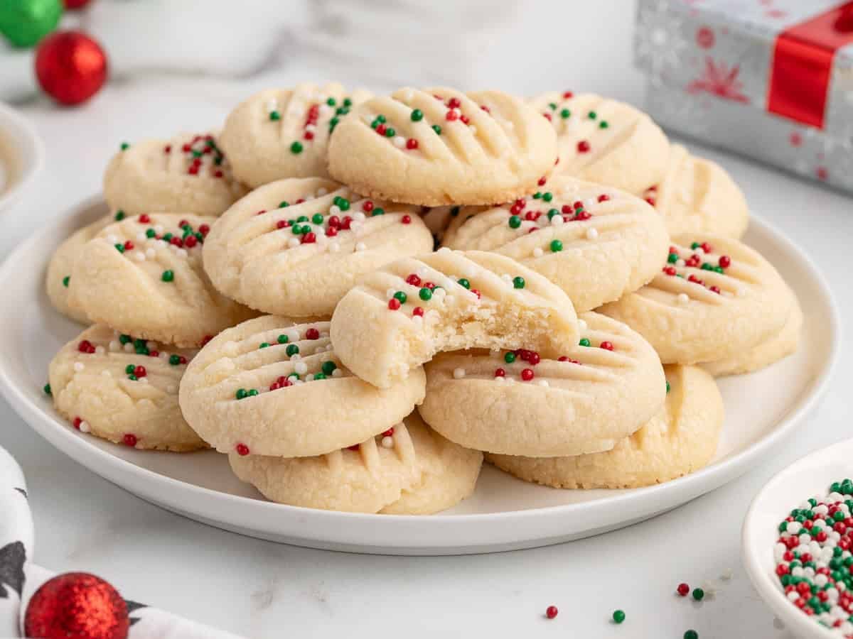 A plate of round, buttery Christmas Shortbread Cookies topped with red, green, and white sprinkles. One cookie is partially eaten. Christmas decorations and a wrapped gift are in the background.