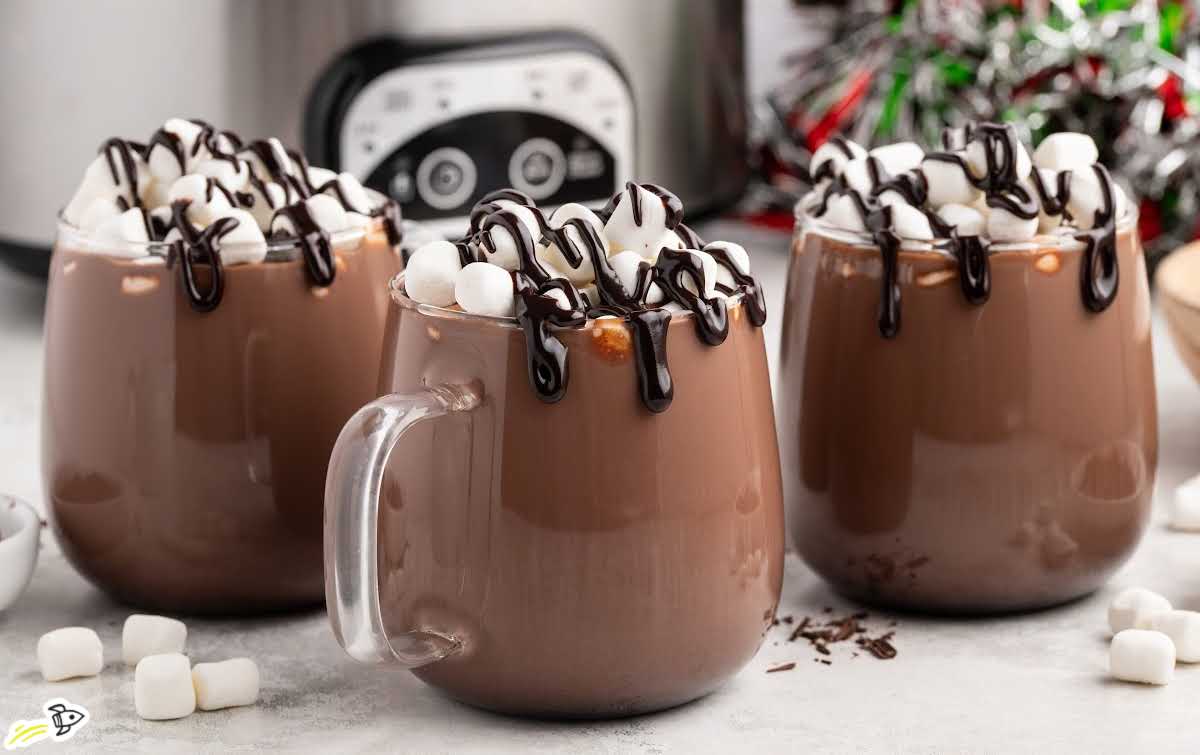 Three clear mugs of crockpot hot chocolate topped with marshmallows and chocolate drizzle in front of a slow cooker.