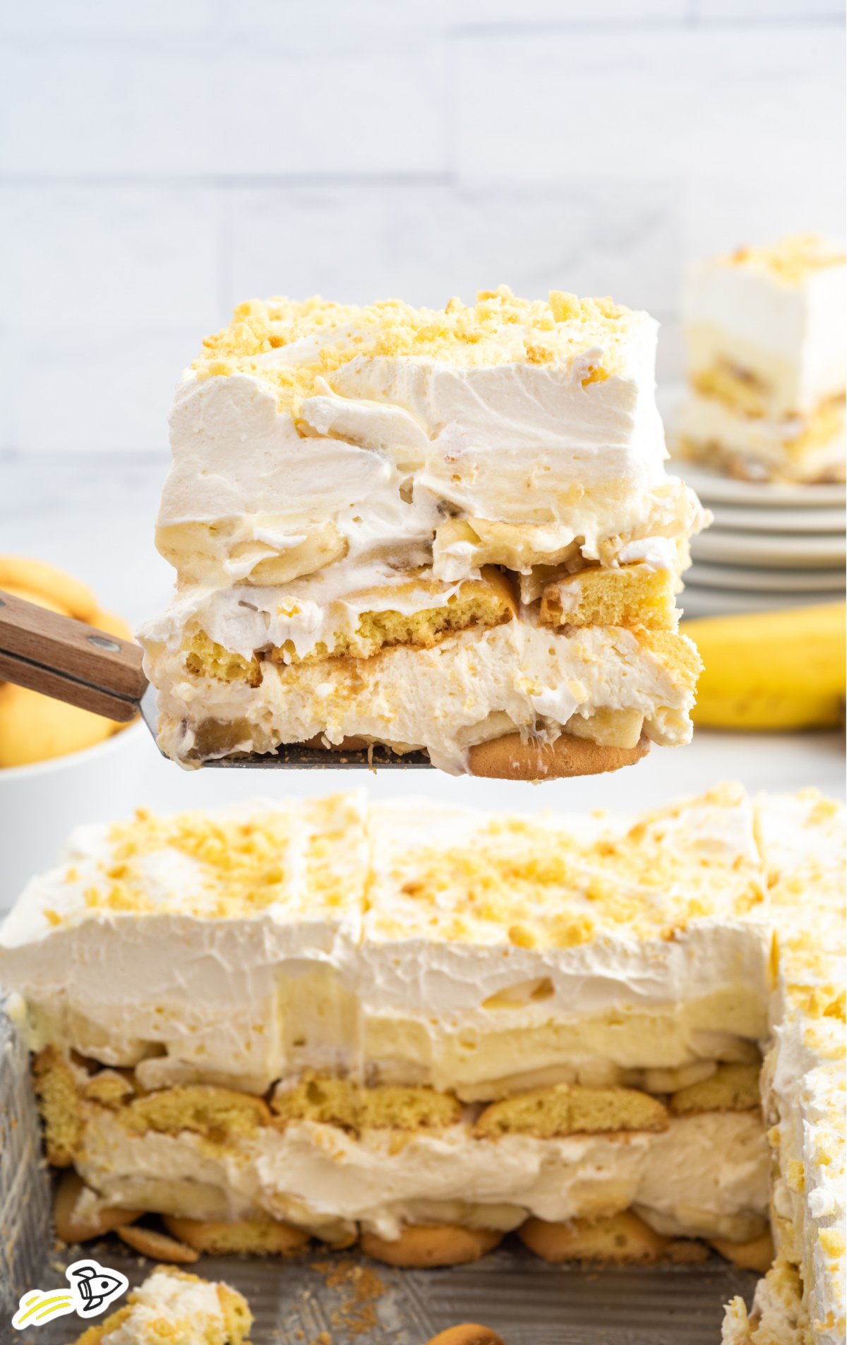 A close-up view of a creamy, layered slice of Banana Delight dessert being lifted from the pan, showing visible layers of vanilla wafers, sliced bananas, creamy filling, and whipped topping, sprinkled with crushed cookie crumbs on top.