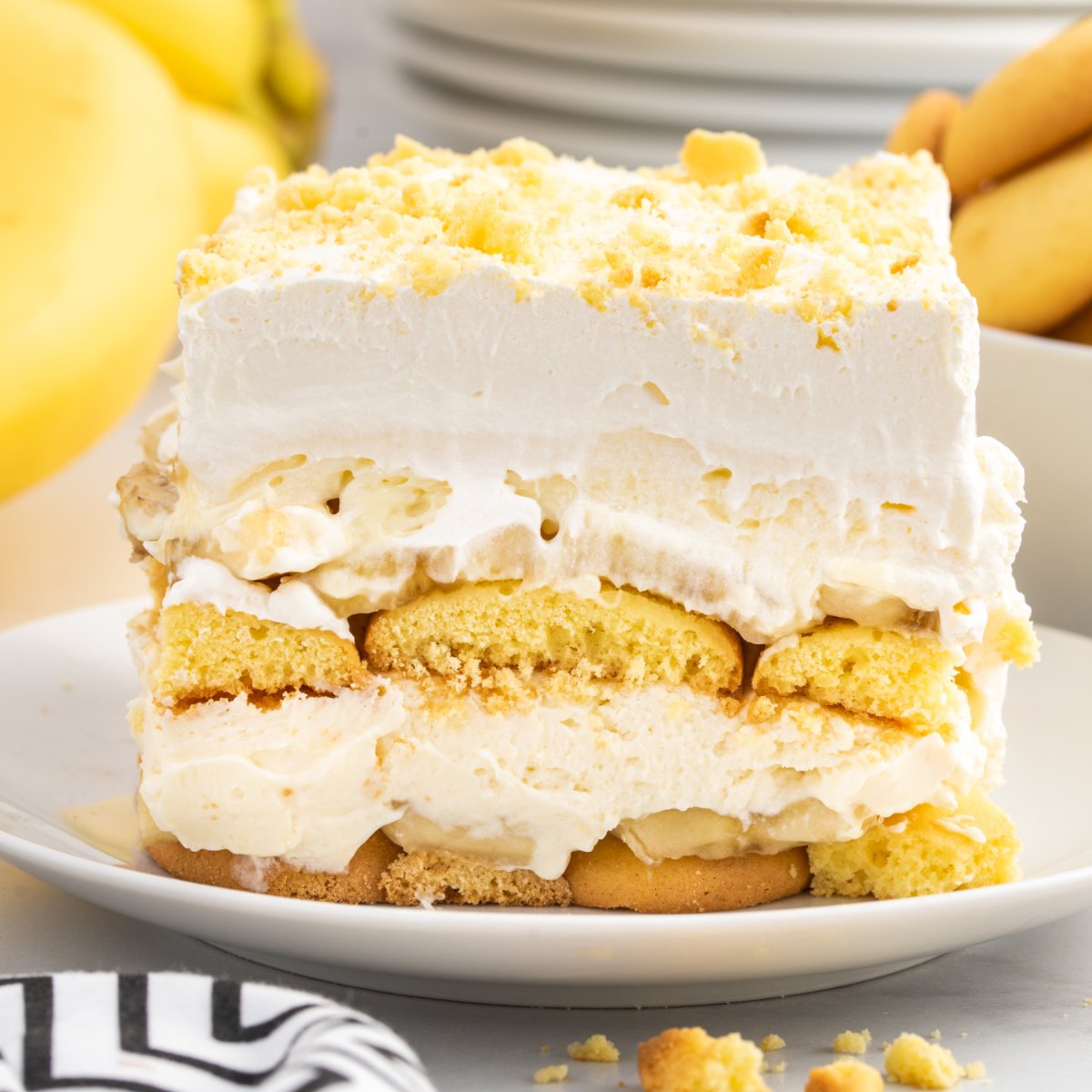 A close-up of a single slice of Banana Delight layered dessert on a white plate, showing thick layers of vanilla wafers, creamy pudding filling, sliced bananas, and whipped topping with crushed cookie crumbs sprinkled on top. Bananas and cookies are visible in the background.