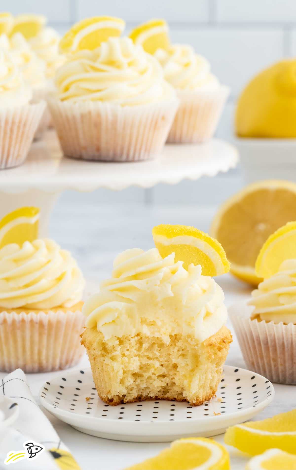 a close up shot of a Lemon Cupcake on a plate topped with a slice of lemon and frosting with a bite taken out of it
