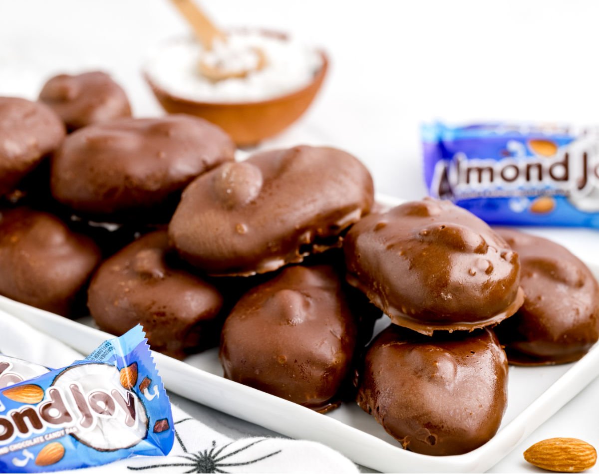 A white platter stacked with homemade Almond Joy bars coated in smooth milk chocolate, with packaged Almond Joy candies and a bowl of shredded coconut in the background.