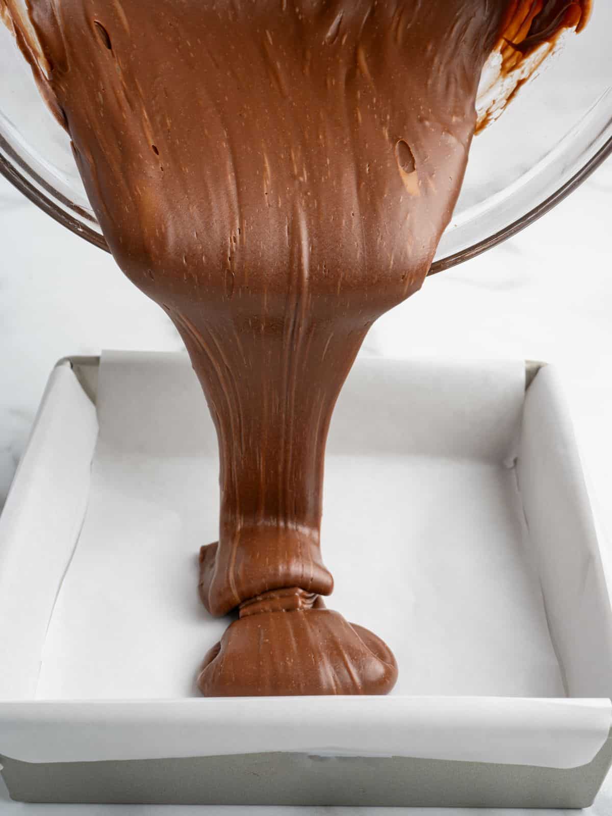 Thick Baileys Fudge chocolate batter being poured from a bowl into a square baking pan lined with parchment paper.
