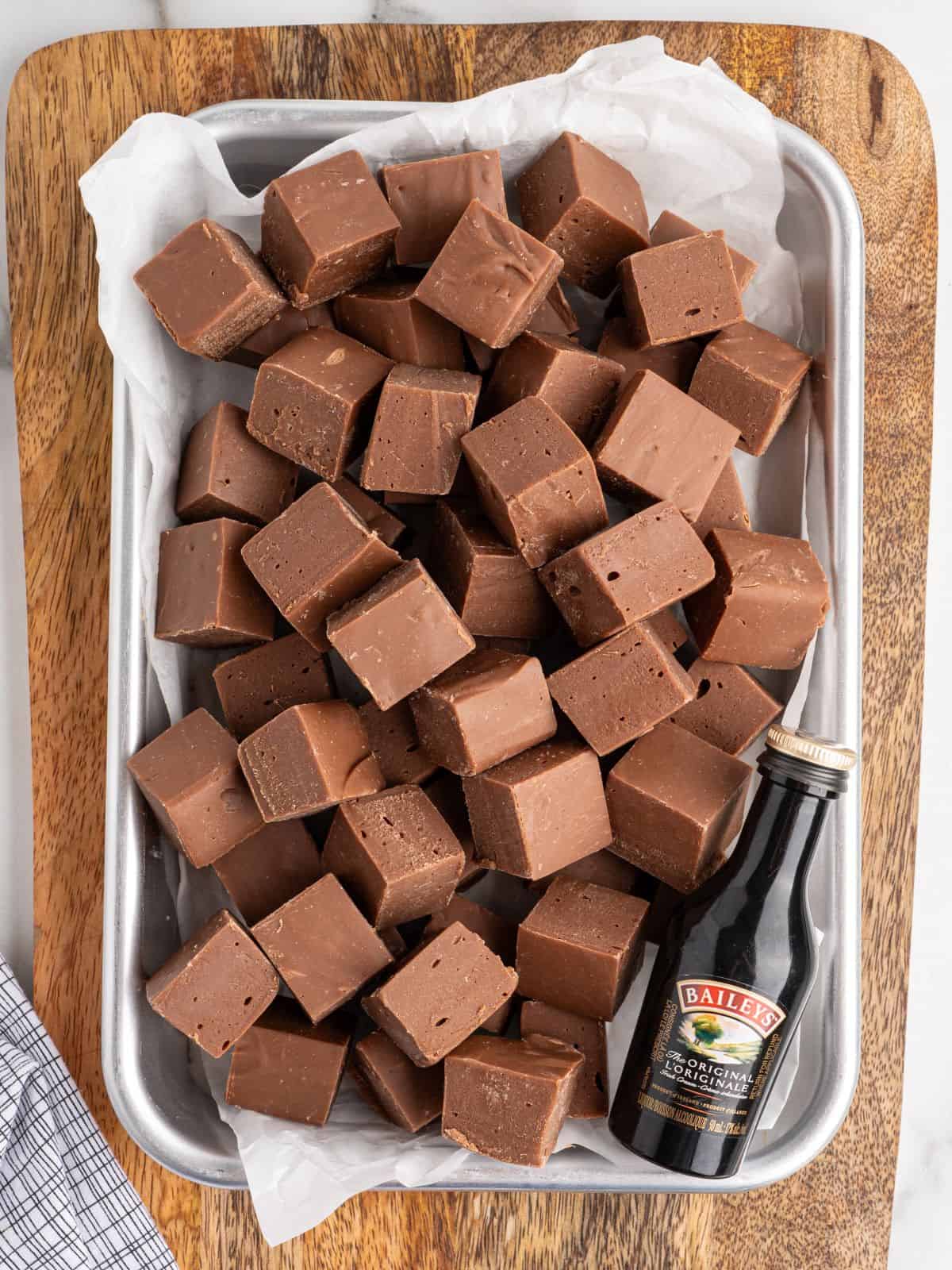 A tray filled with square pieces of Baileys Fudge sits on a wooden board, accompanied by a small bottle of Baileys Irish Cream in the corner.