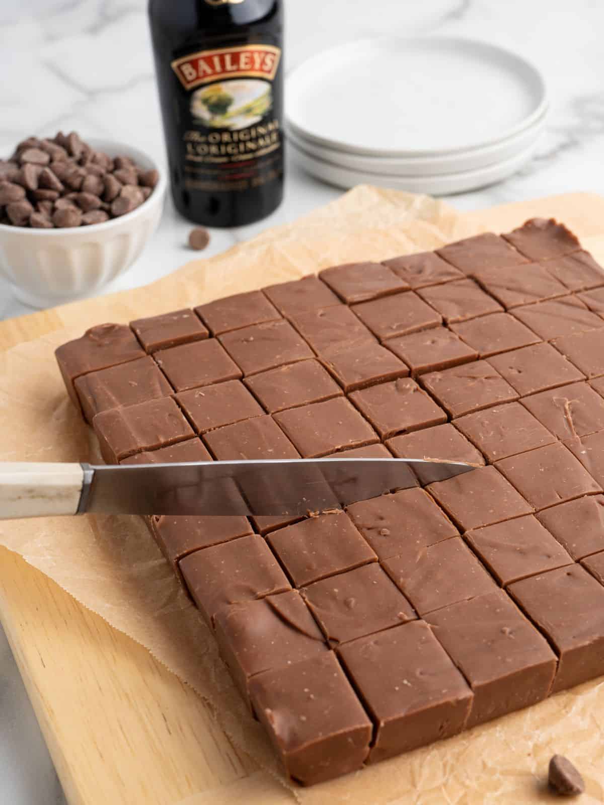 A large block of Baileys Fudge cut into squares sits on parchment paper, with a knife mid-slice. In the background are a bottle of Baileys, a bowl of chocolate chips, and stacked white plates.