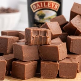 A pile of creamy Baileys Fudge squares, with one piece showing bite marks, sits in front of a blurred bottle of Baileys Irish Cream.