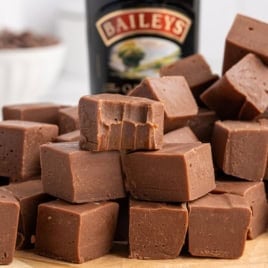 A stack of creamy Baileys Fudge cubes, with one piece showing bite marks, sits in front of a blurred bottle of Baileys Irish Cream.