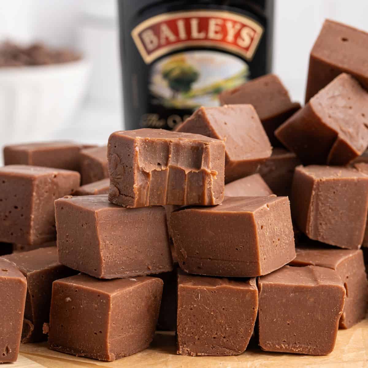 A stack of creamy Baileys Fudge cubes, with one piece showing bite marks, sits in front of a blurred bottle of Baileys Irish Cream.