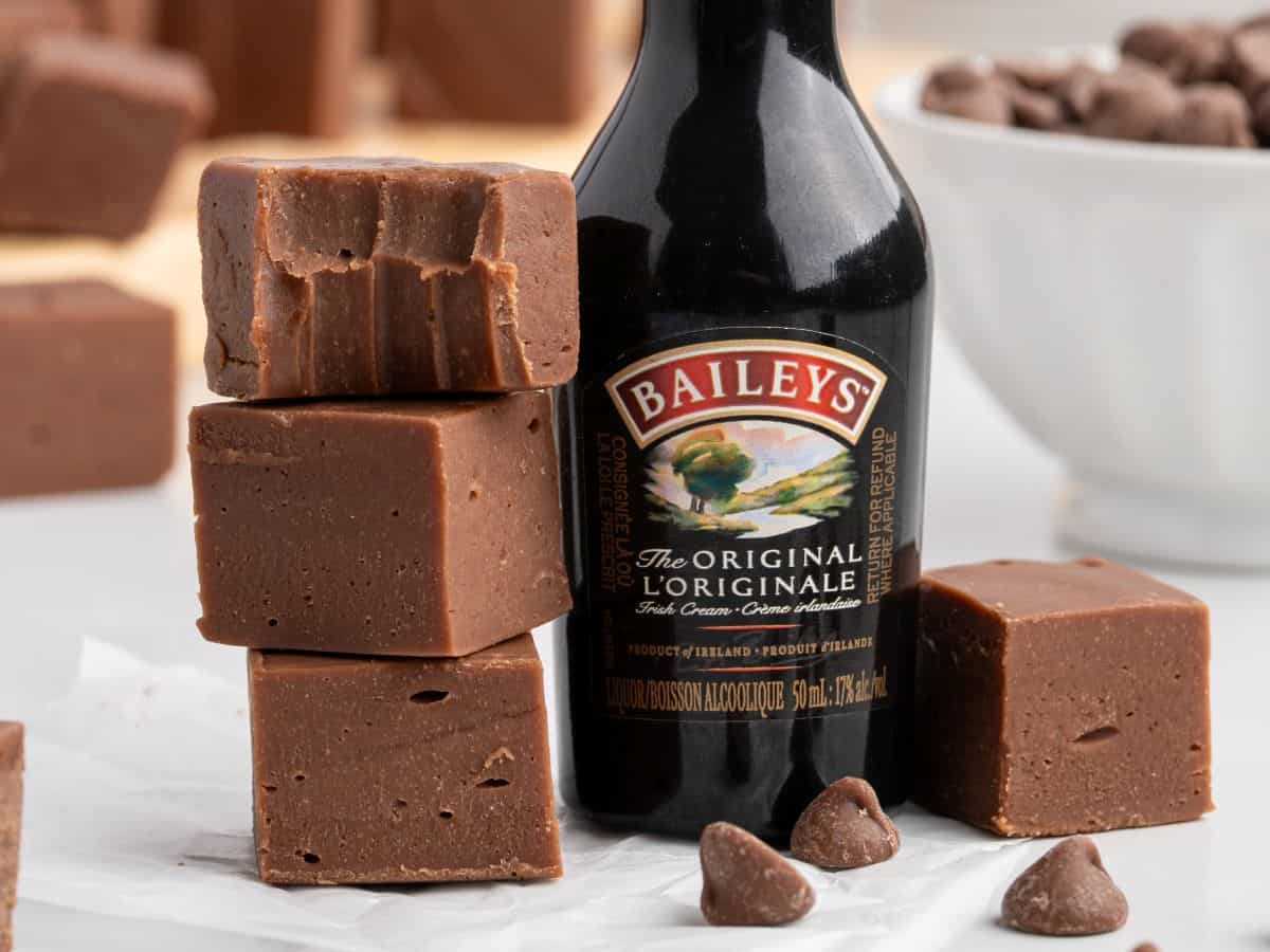A stack of rich Baileys Fudge squares sits next to a bottle of Baileys Irish Cream, with more fudge and chocolate chips scattered in the background on a white surface.