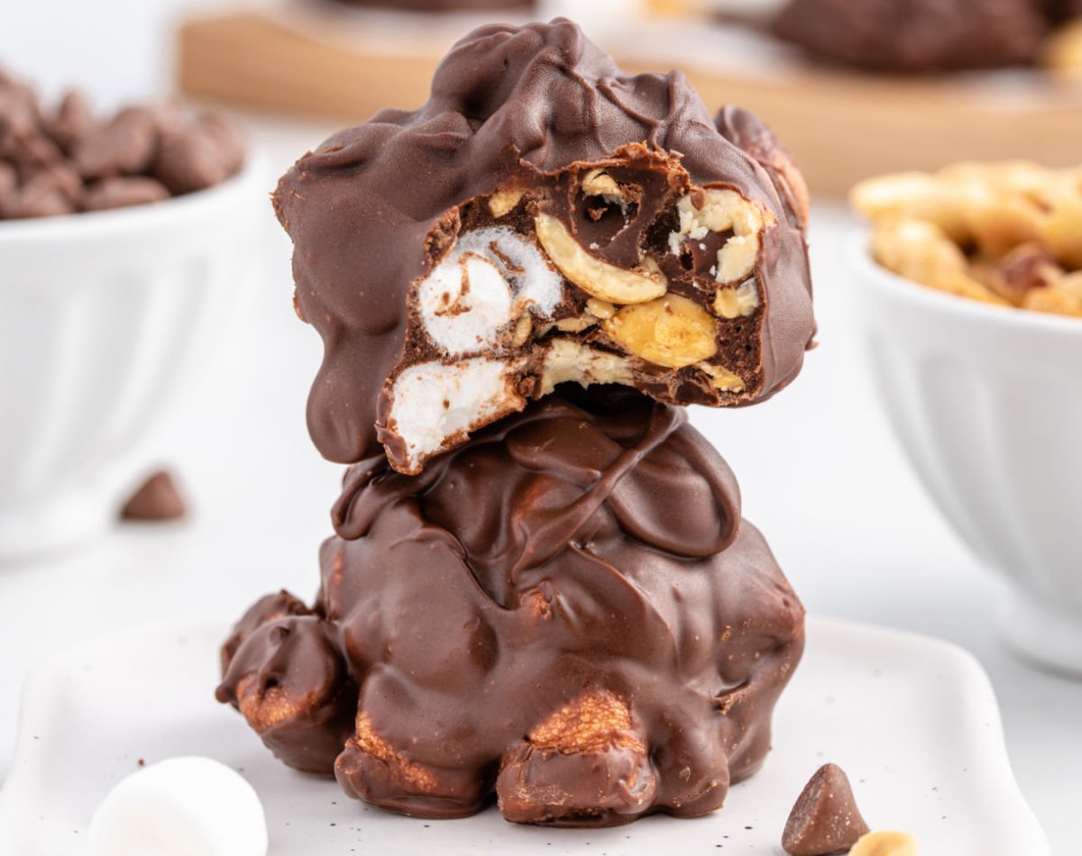 A close-up of two stacked peanut marshmallow clusters coated in glossy chocolate. The top cluster is broken open to reveal whole mini marshmallows, crunchy peanuts, and thick chocolate layers inside, with bowls of chocolate chips and peanuts blurred in the background.
