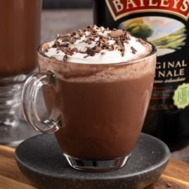 Steaming mug of Baileys hot chocolate with whipped cream and chocolate curls beside a bottle of Baileys.