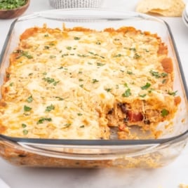 A glass baking dish filled with a cheesy Chicken Taco Casserole, topped with chopped herbs. A portion has been served, revealing flavorful layers of chicken, zesty sauce, and melted cheese inside.