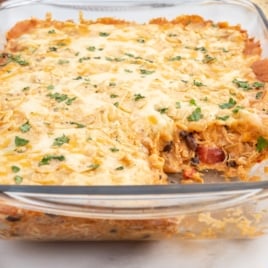 A glass baking dish filled with a cheesy Chicken Taco Casserole, topped with melted cheese and chopped herbs, with one serving scooped out from the corner.
