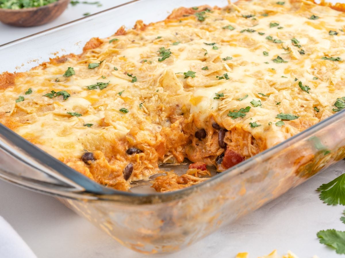A baked Chicken Taco Casserole in a glass dish, topped with melted cheese, chopped herbs, and bits of tortilla chips, with a serving removed to reveal layers of beans, sauce, and tender chicken inside.