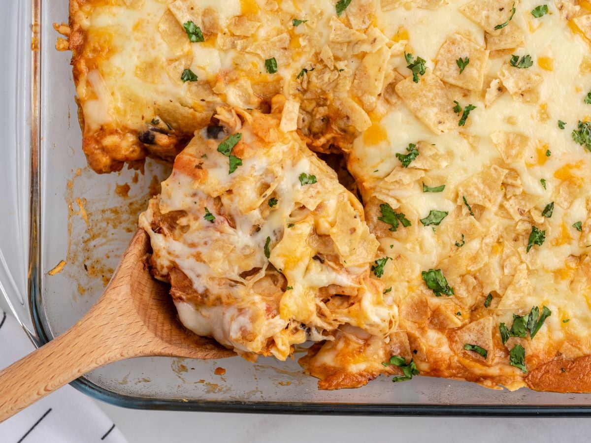 A wooden spoon serves a portion of cheesy Chicken Taco Casserole topped with crushed tortilla chips and chopped parsley from a glass baking dish.