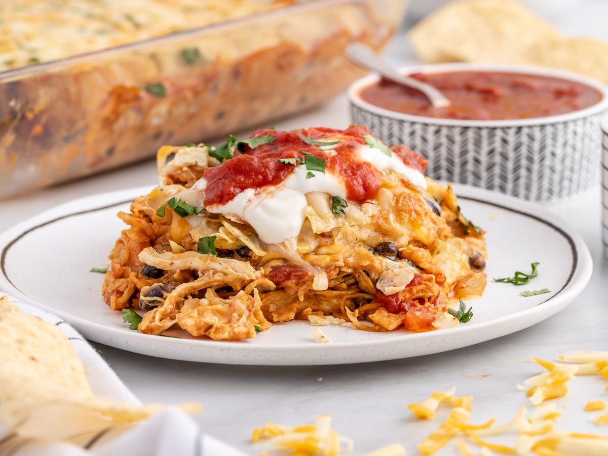 A serving of cheesy Chicken Taco Casserole topped with sour cream, salsa, and cilantro sits on a white plate. In the background, there’s a bowl of salsa and the rest of the casserole in a baking dish.