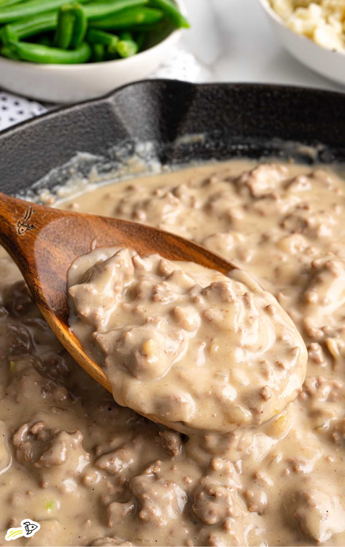Wooden spoon scooping thick hamburger gravy from a cast iron skillet, with a bowl of green beans in the background.