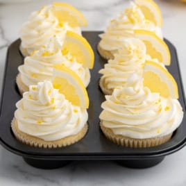 Six Lemon Cupcakes in a muffin tin, topped with swirls of white frosting, lemon zest, and garnished with a slice of lemon on each, set on a marble surface.