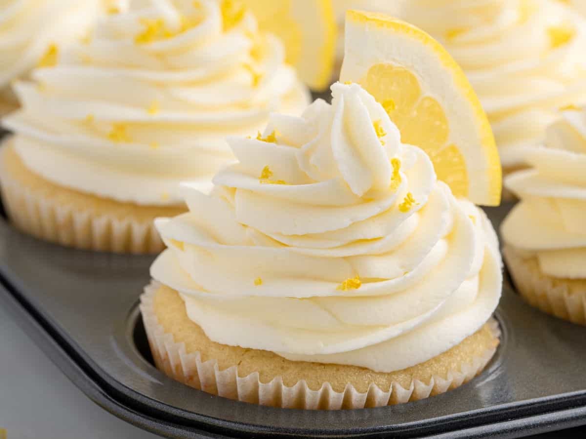 A close-up of Lemon Cupcakes with swirled white frosting, topped with lemon zest and a slice of lemon, sitting in a muffin tin with other similarly decorated cupcakes in the background.