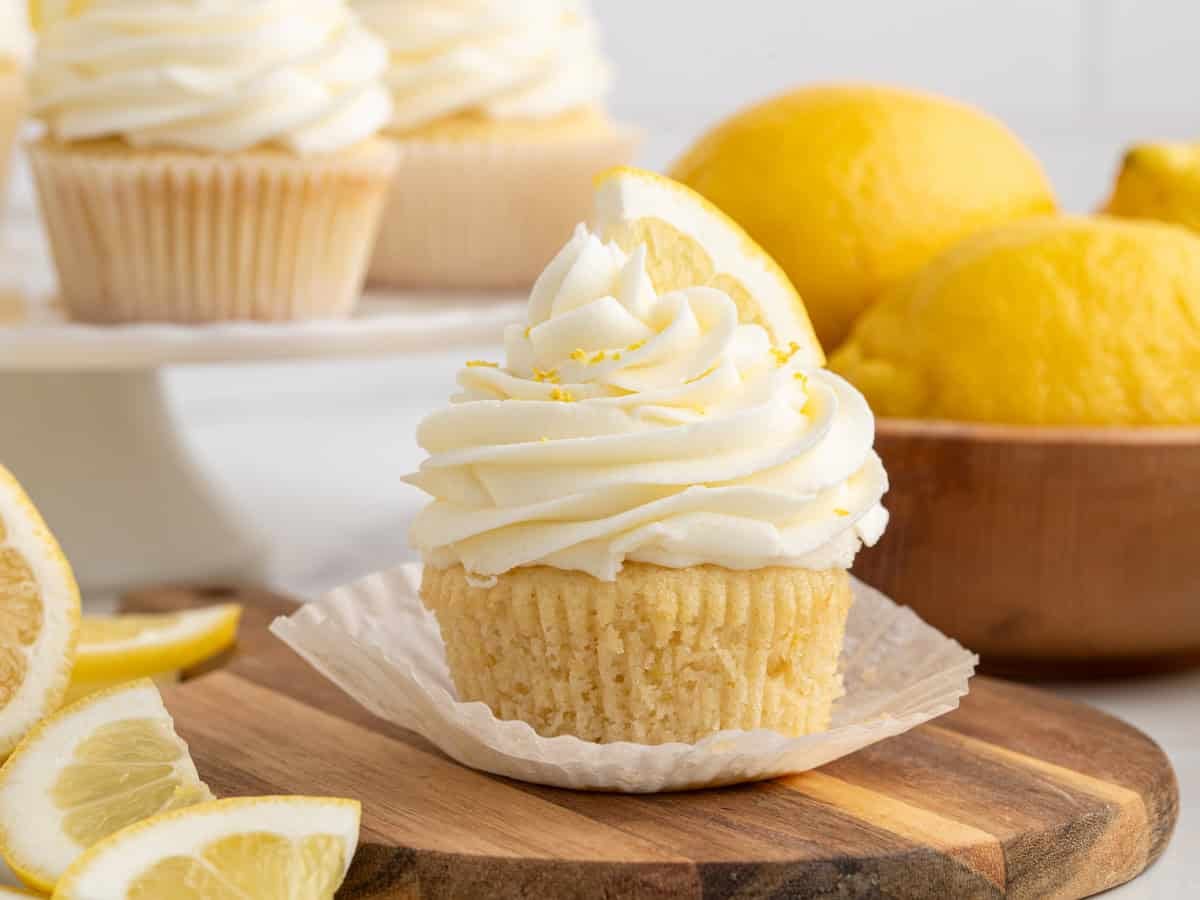 A Lemon Cupcakes delight with swirled white frosting and a slice of lemon on top sits on a wooden board, surrounded by whole lemons and more lemon cupcakes in the background.