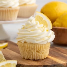 A Lemon Cupcakes masterpiece: a lemon cupcake with swirled white frosting and a lemon slice on top sits on a wooden board, surrounded by whole lemons and more cupcakes in the background.