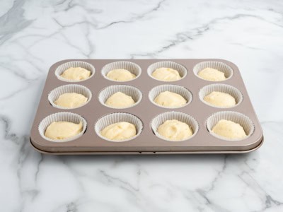 A 12-cup muffin tin lined with white paper liners, each filled with unbaked Lemon Cupcakes batter, sits on a white marble countertop.