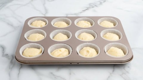 A 12-cup muffin tin lined with white paper liners, each filled with unbaked Lemon Cupcakes batter, sits on a white marble countertop.