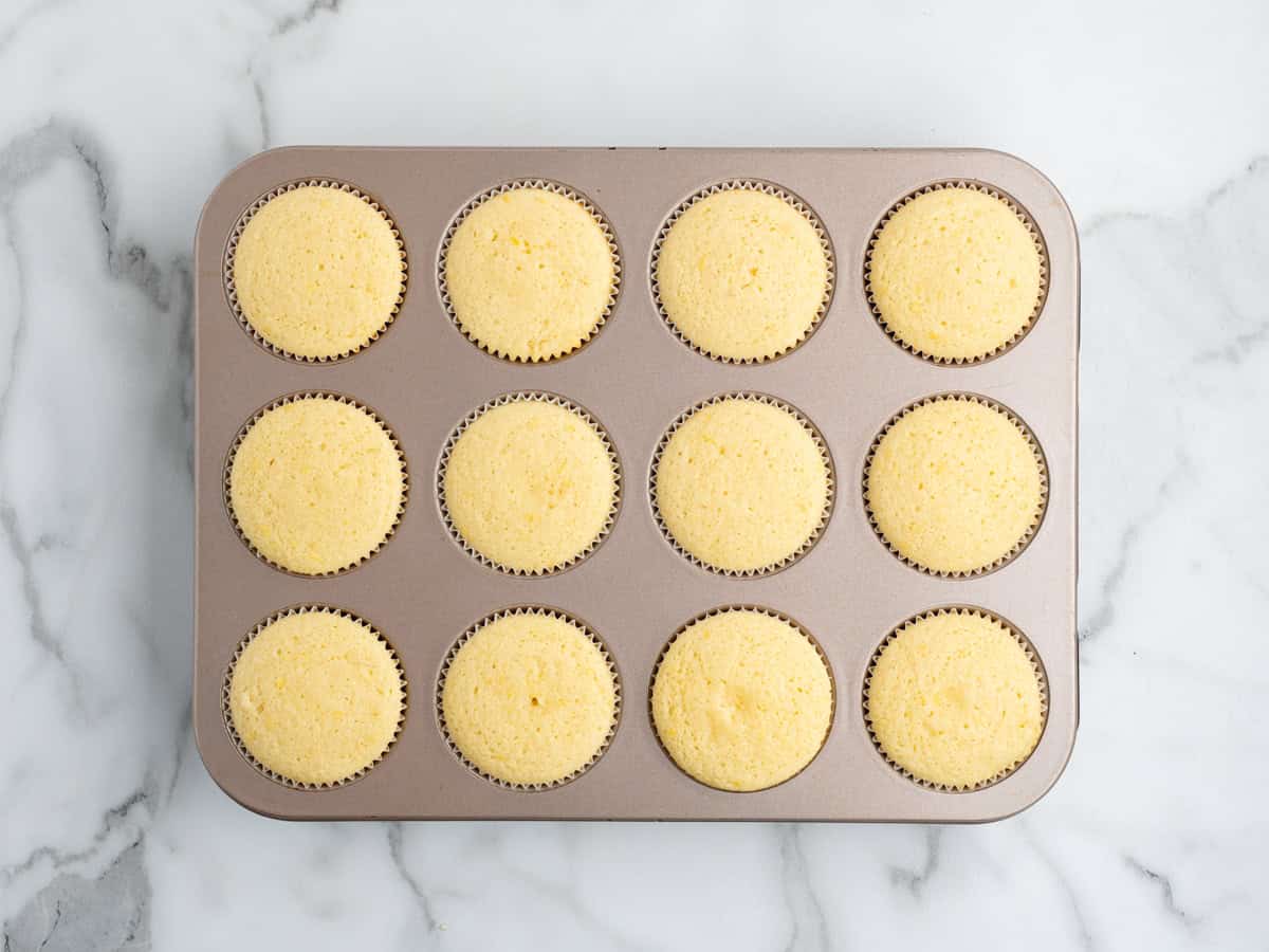 A metal muffin tin holds twelve freshly baked lemon cupcakes in white paper liners, resting on a white marble surface.