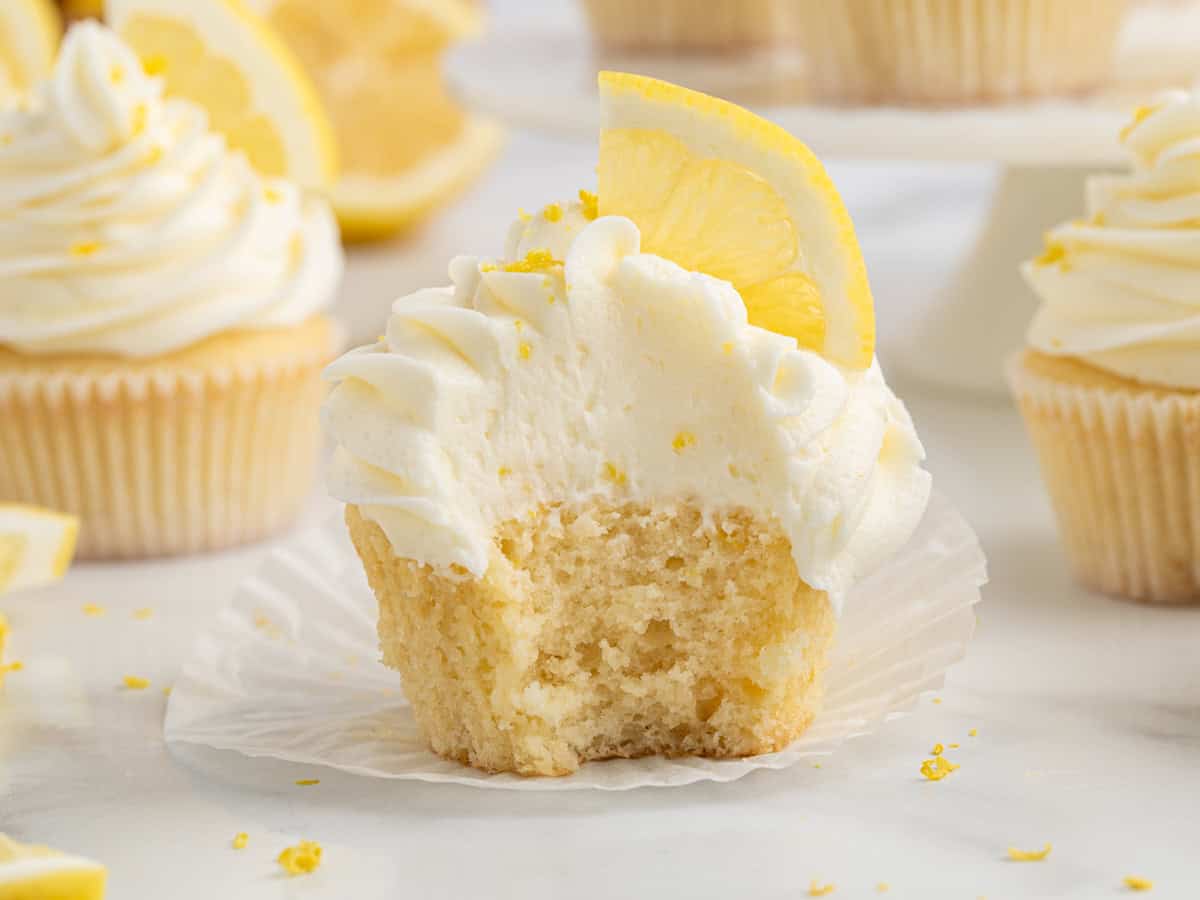 A Lemon Cupcakes delight with white frosting and a lemon slice on top, partially unwrapped with a large bite taken out, sits on a white surface surrounded by other cupcakes and lemon pieces in the background.