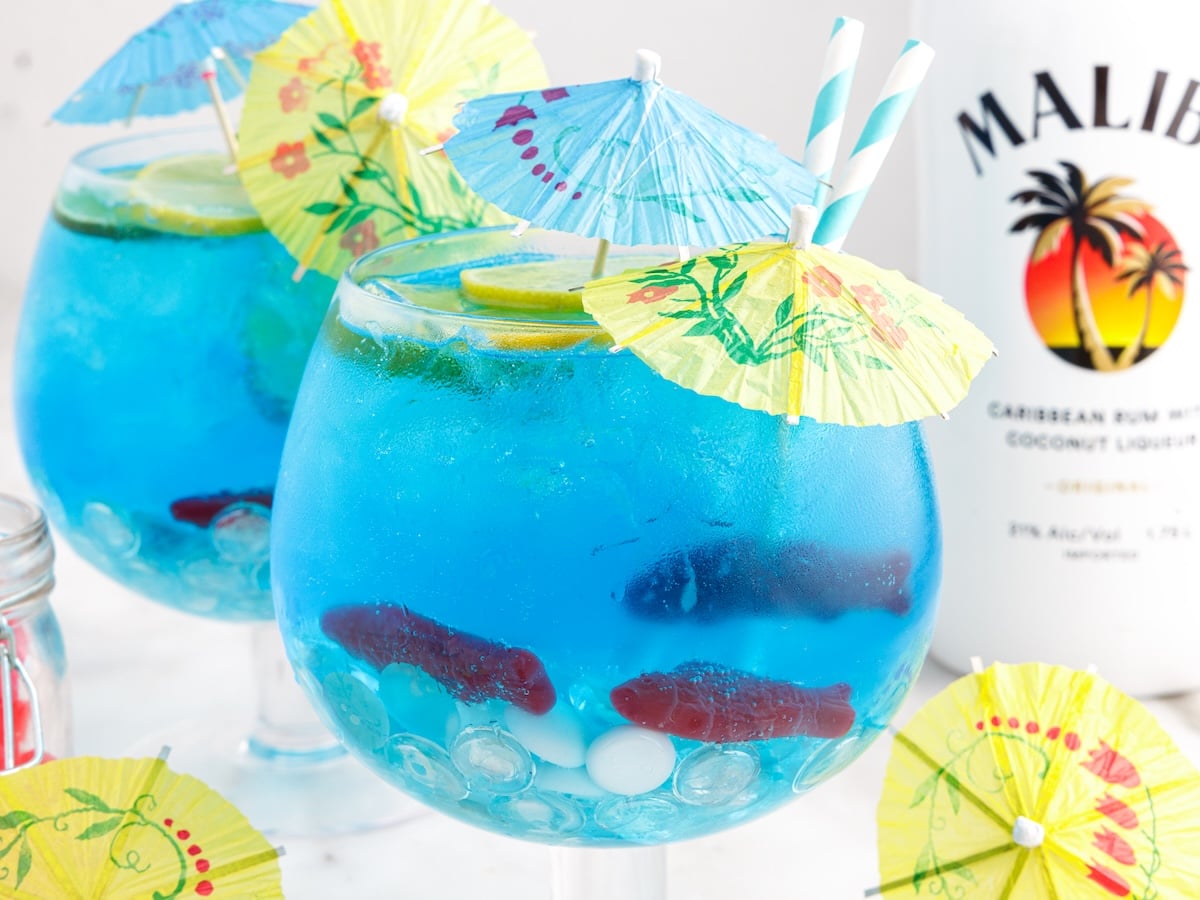 Two large round glasses filled with bright blue Fish Bowl Drink cocktails, ice, lime slices, and gummy fish, each topped with colorful paper umbrellas and striped straws. A bottle of Malibu rum is in the background.