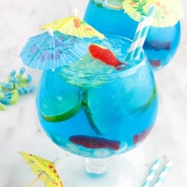 A large glass of bright blue Fish Bowl Drink with ice, lime slices, gummy candies, a striped straw, and a colorful paper umbrella, with another similar drink and extra umbrellas in the background.