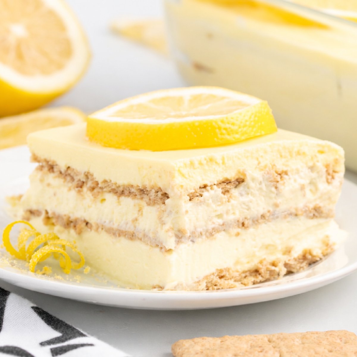 Slice of lemon eclair cake on a white plate, showing creamy lemon pudding layers and softened graham crackers, topped with a fresh lemon slice and garnished with thick lemon zest curls, with whole lemons and the baking dish in the background.