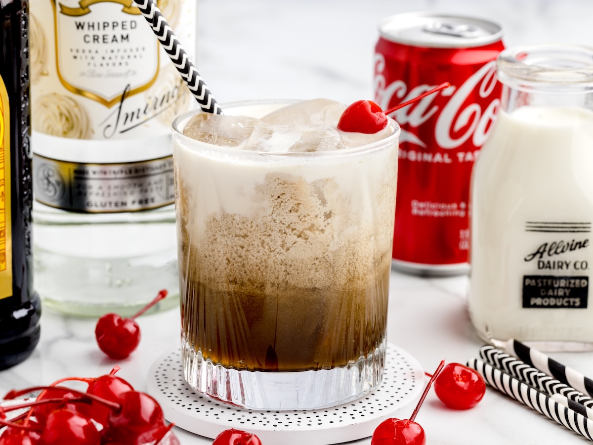 A glass of Paralyzer Drink with cream, cola, and a cherry on top sits on a coaster. Surrounding it are bottles of whipped cream vodka, cola, milk, and maraschino cherries. A striped straw is in the glass.