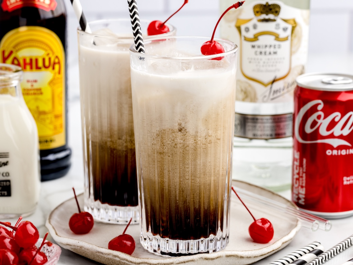Two tall glasses of Paralyzer Drink filled with a creamy brown cocktail topped with foam and red cherries, surrounded by bottles of Kahlua, whipped cream vodka, milk, a can of Coca-Cola, and extra cherries on a white plate.
