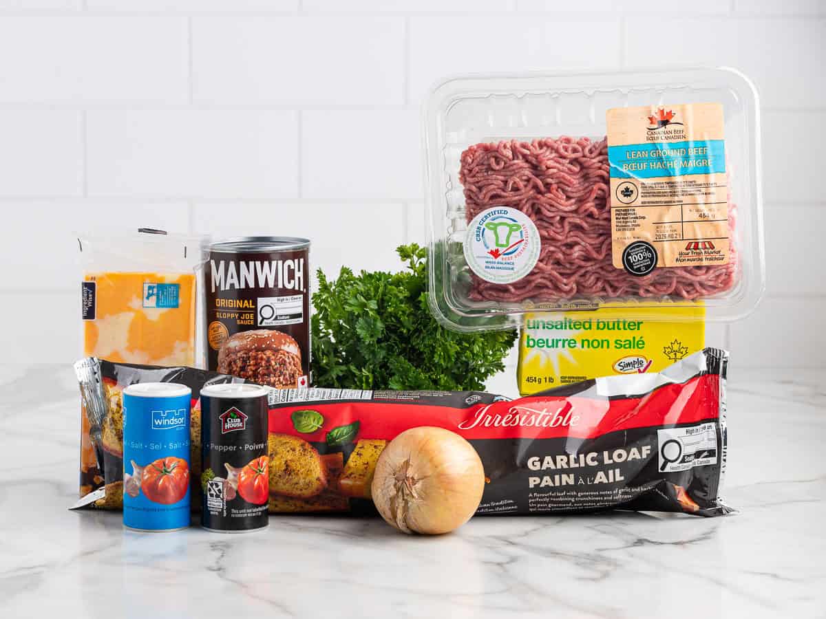 Assorted ingredients on a counter—ground beef, parsley, onion, butter, cheddar cheese, garlic loaf, canned tomatoes, Manwich sauce, and salt—all set against a white tile background for preparing delicious Garlic Bread Sloppy Joes.