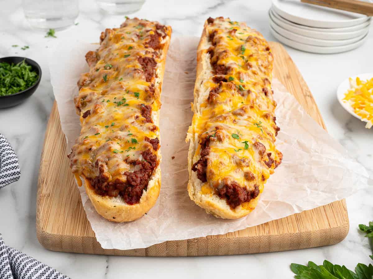 Two cheesy French bread pizzas, inspired by Garlic Bread Sloppy Joes, are topped with savory ground beef and fresh parsley and sit on parchment paper over a wooden board, with plates, cheese, and herbs in the background.