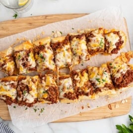 Sliced French bread topped with seasoned ground beef and melted cheese, served on parchment paper atop a wooden board, with parsley and shredded cheese nearbyโa delicious twist on Garlic Bread Sloppy Joes.