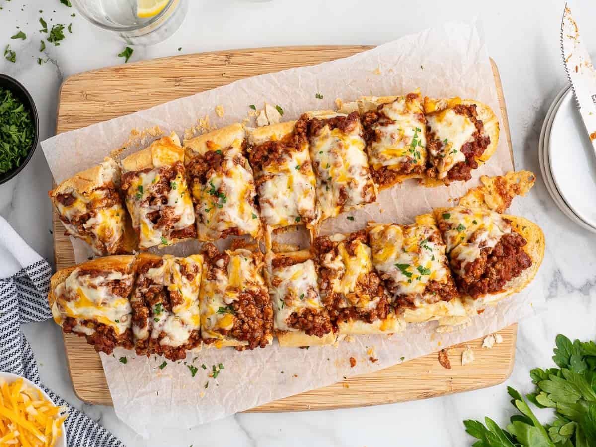 Sliced French bread topped with seasoned ground beef and melted cheese, served on parchment paper atop a wooden board, with parsley and shredded cheese nearby—a delicious twist on Garlic Bread Sloppy Joes.