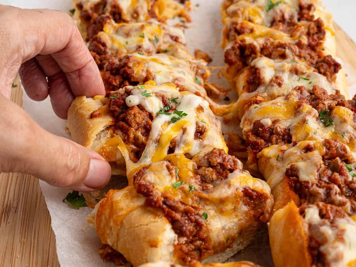 A hand picks up a cheesy, baked Garlic Bread Sloppy Joes bread topped with ground meat, melted cheese, and herbs. Several more pieces are arranged on parchment paper on a wooden surface.