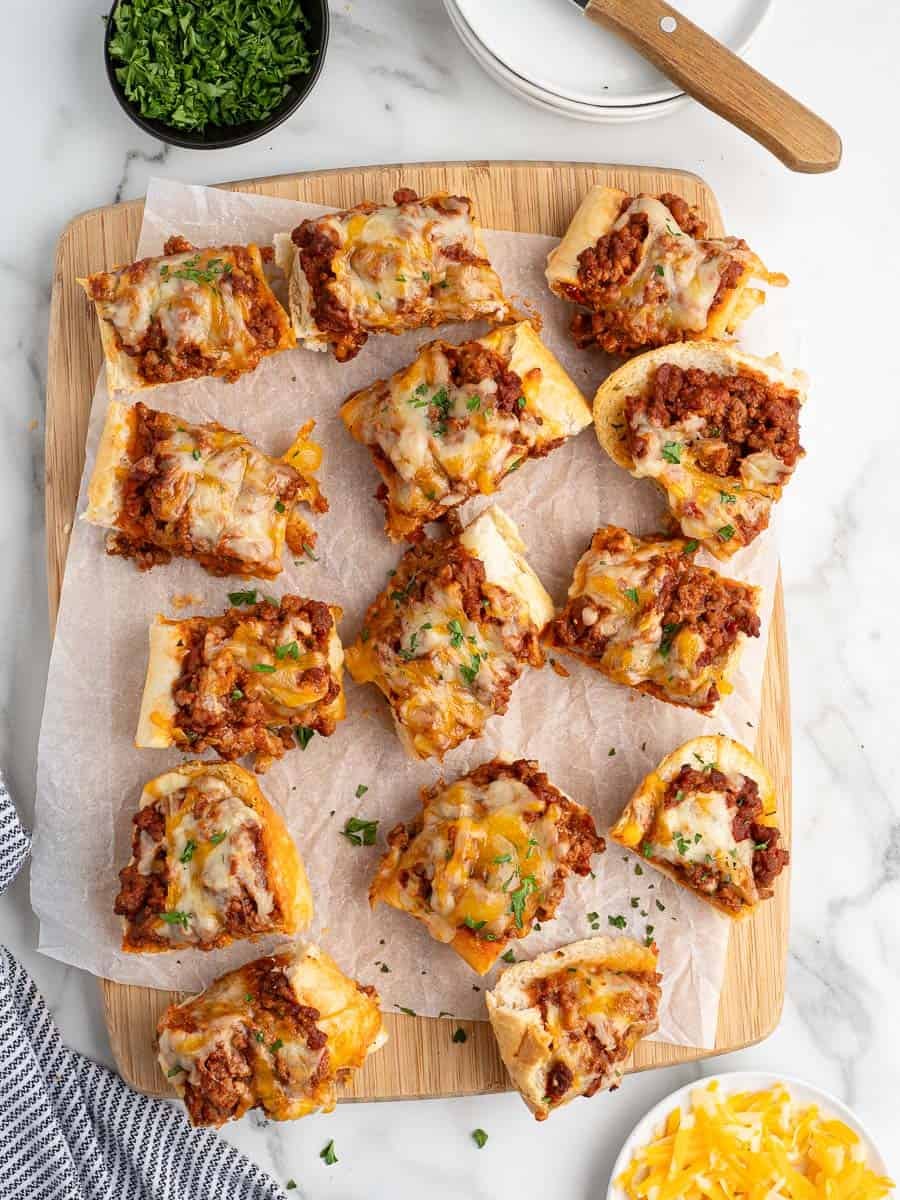 Several pieces of cheesy ground beef French bread pizza—reminiscent of Garlic Bread Sloppy Joes—are arranged on a wooden board lined with parchment paper. Chopped herbs, plates, and shredded cheese are nearby.