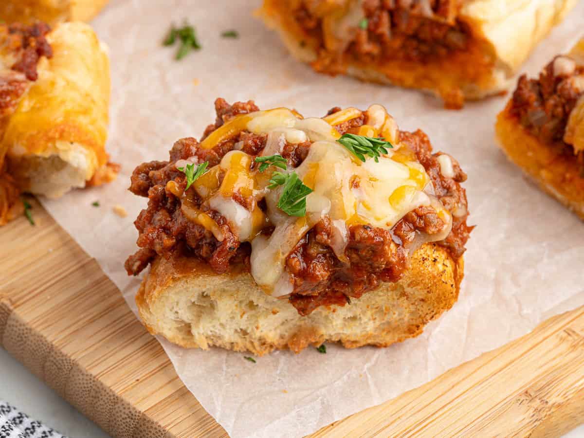Toasted garlic bread topped with saucy ground beef, melted cheese, and a sprinkle of fresh parsley sits on parchment paper on a wooden board—an irresistible twist on classic Garlic Bread Sloppy Joes.