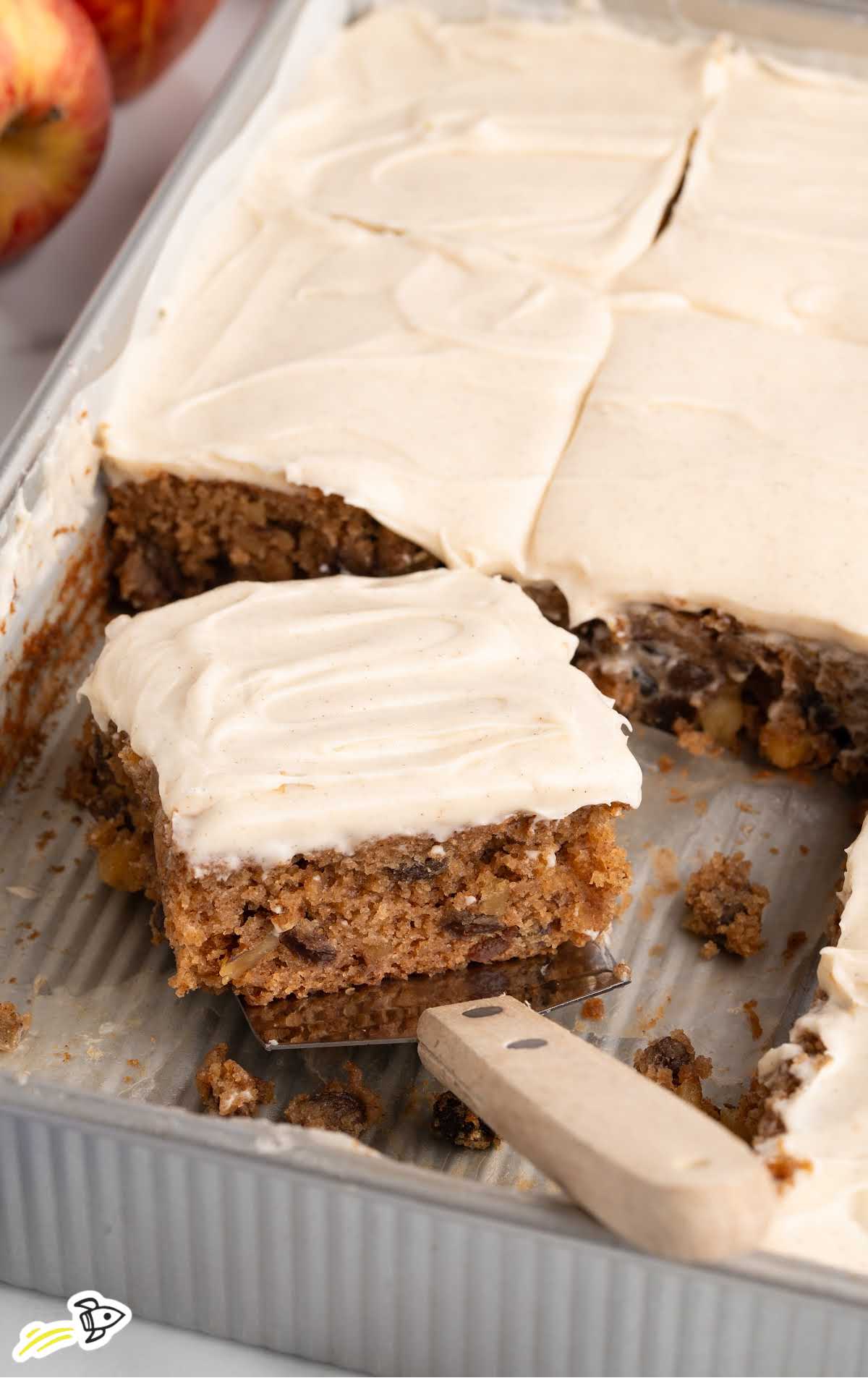 Pan of applesauce snack cake with cream cheese frosting, one square being lifted with a spatula