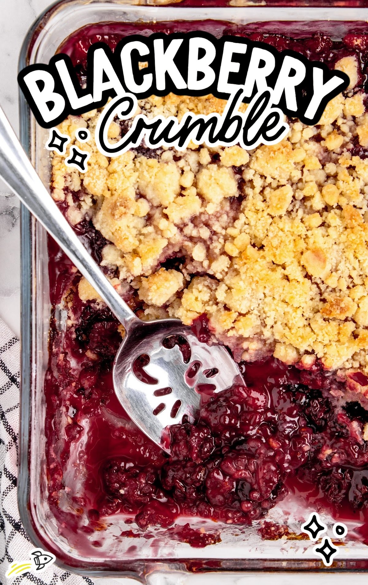 Overhead close-up of a glass baking dish filled with juicy blackberry crumble, showing golden-brown crumb topping and glossy baked blackberries, with a silver serving spoon resting in the dish.