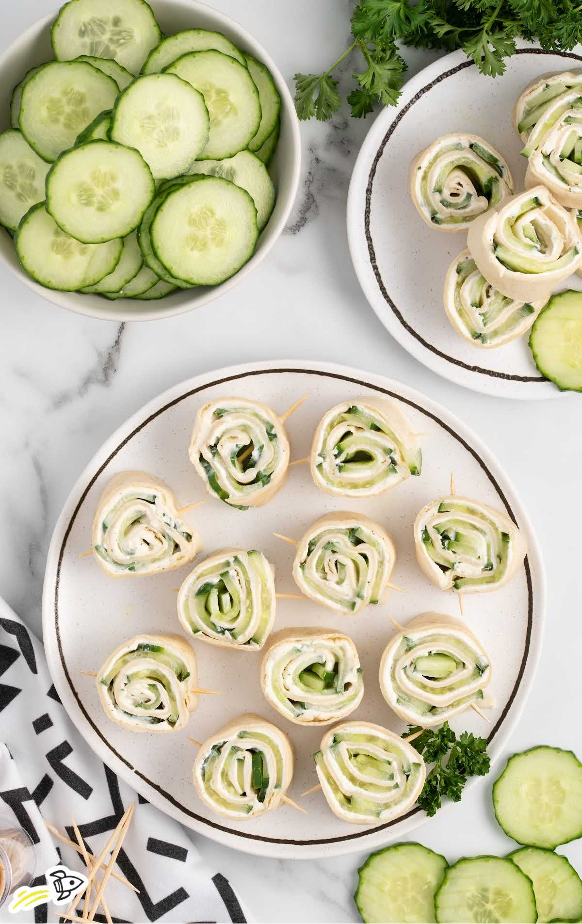 overhead shot of plates of Pinwheel Sandwiches with a bowl of sliced cucumbers