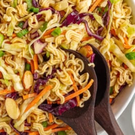 A close-up of a vibrant Ramen Noodle Salad featuring ramen noodles, shredded cabbage, carrots, green onions, and sliced almonds, all tossed together in a large white bowl with wooden salad servers.