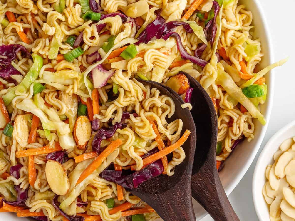 A close-up of a vibrant Ramen Noodle Salad featuring ramen noodles, shredded cabbage, carrots, green onions, and sliced almonds, all tossed together in a large white bowl with wooden salad servers.